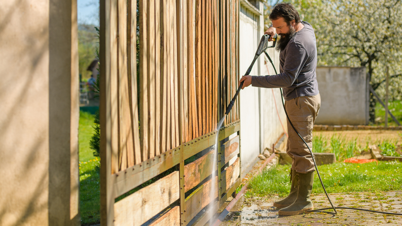 Mid adult man cleaning a wooden gate with a power washer. High pressure water cleaner used to DIY repair garden gate.