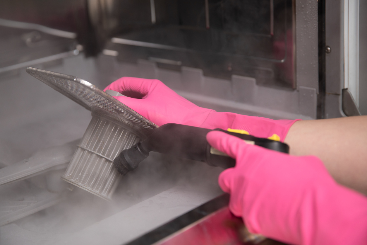Hands in rubber gloves cleaning dishwasher filter with a hot steam cleaner.