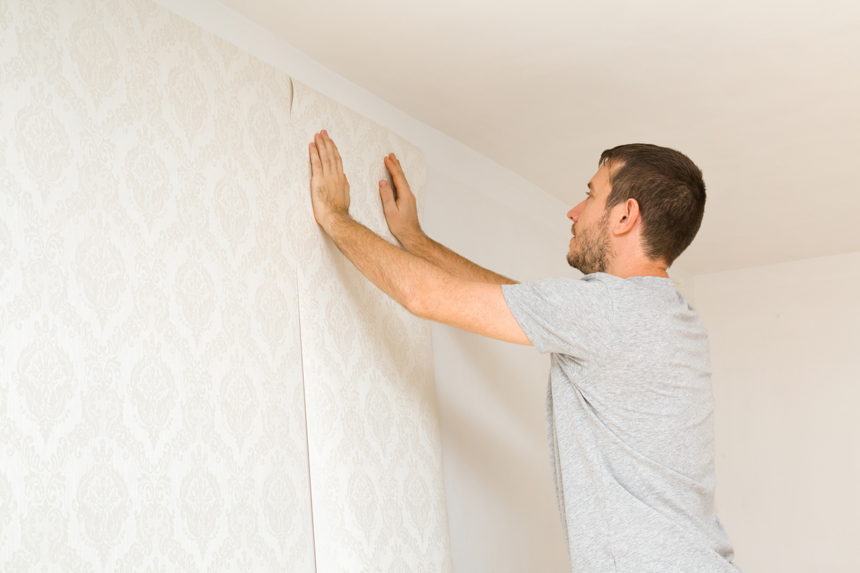 Young adult man hands applying new wallpaper on white wall. Closeup. Repair work of home. Side view.