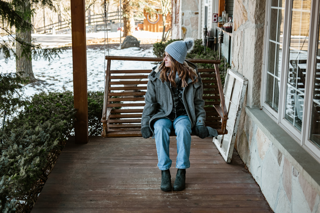 A young woman in winter clothes sits on a porch swing looking out onto the snow covered yard.
