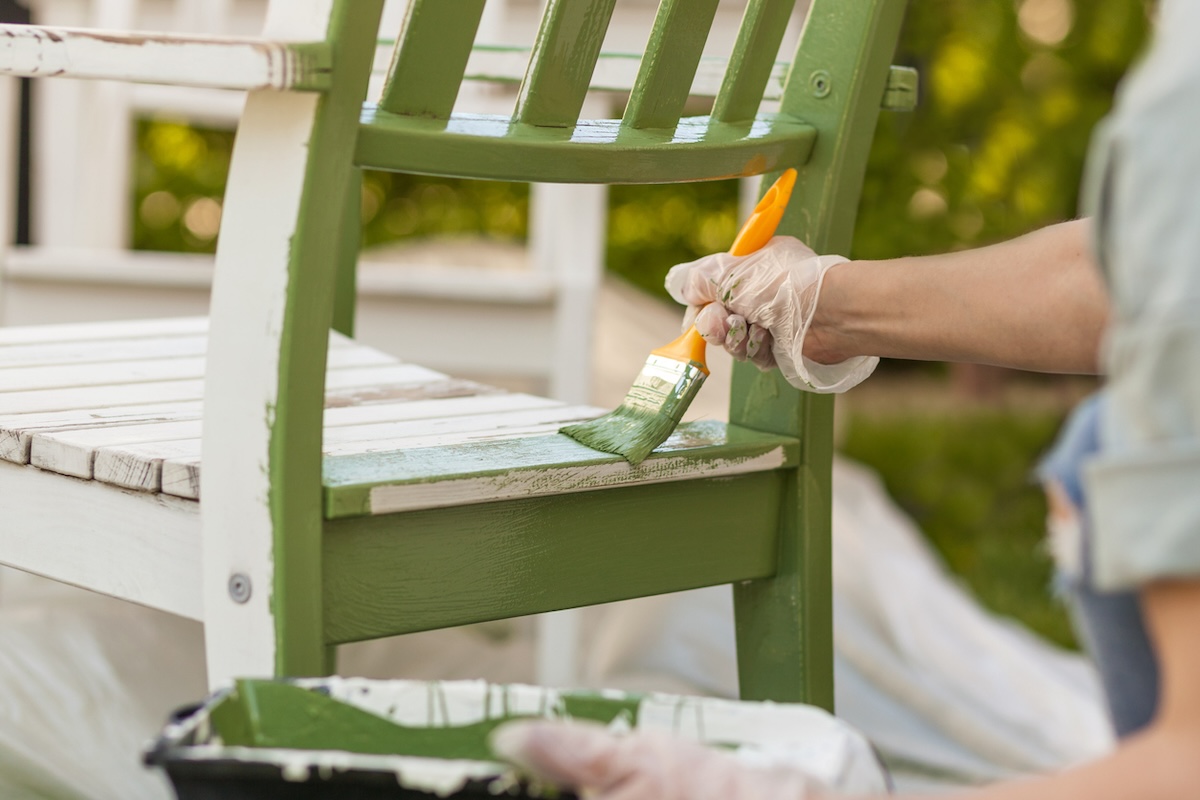 A person painting a wooden patio chair with green exterior furniture paint.