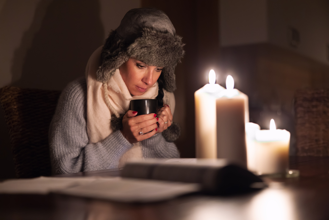 Freezing young woman in winter clothes warms her hands on cup of tea and lights with candles as energy blackouts cause electricity outages.