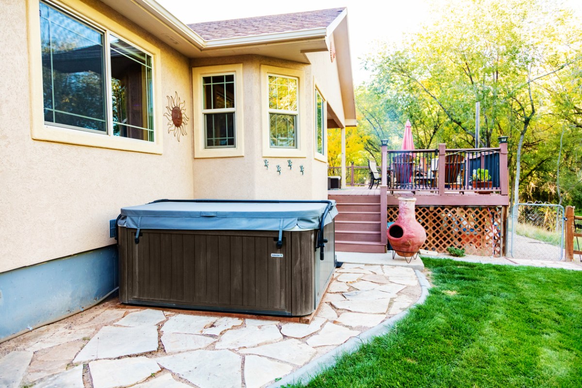 Aging hot tub sitting on stone patio in sunny backyard of a tan home.