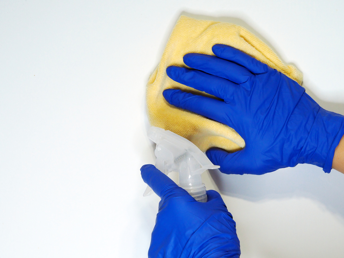 A hand in a protective blue glove cleans the white surface of a cabinet or wall with a white vinegar. Spring cleaning concept.