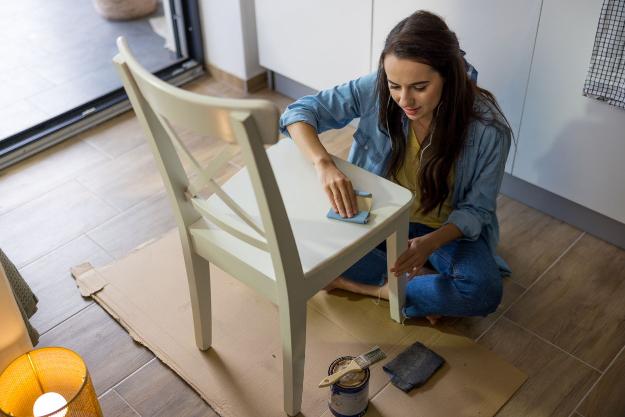 Young female with long brown hair is grinding the top of a white wooden chair with sandpaper,sitting on the ground indoors during daylight,listening to music with her white earphones,paint can with paint brush standing on the ground next to the chair,kitchen in the background