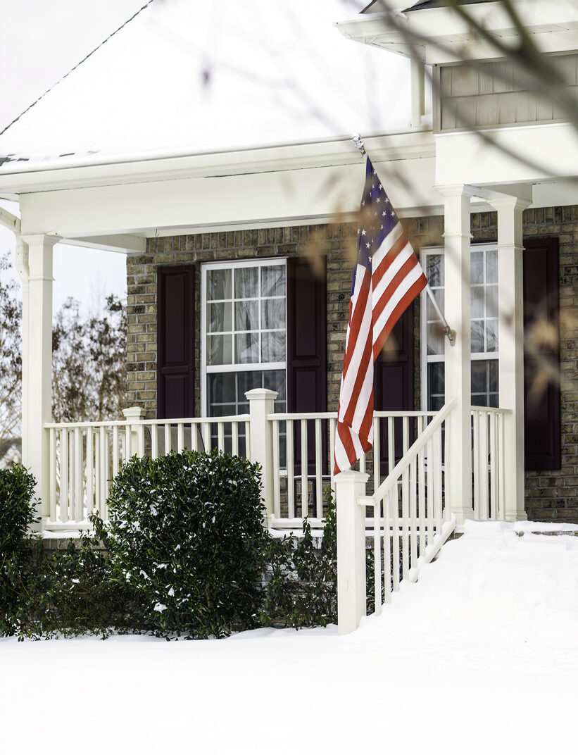 A front porch covered in snow with an American flag hanging out front.