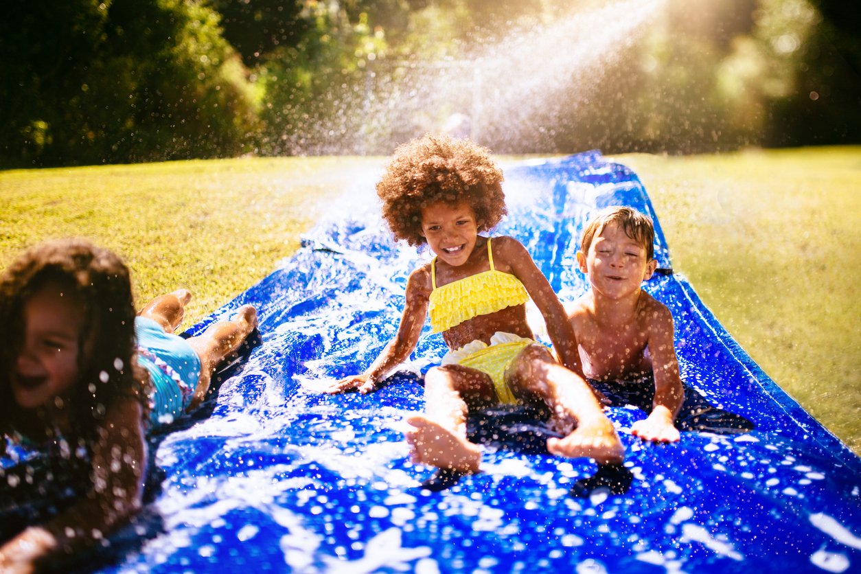 Laughing Black girl and friends playing together on a water slide in their yard in the sun