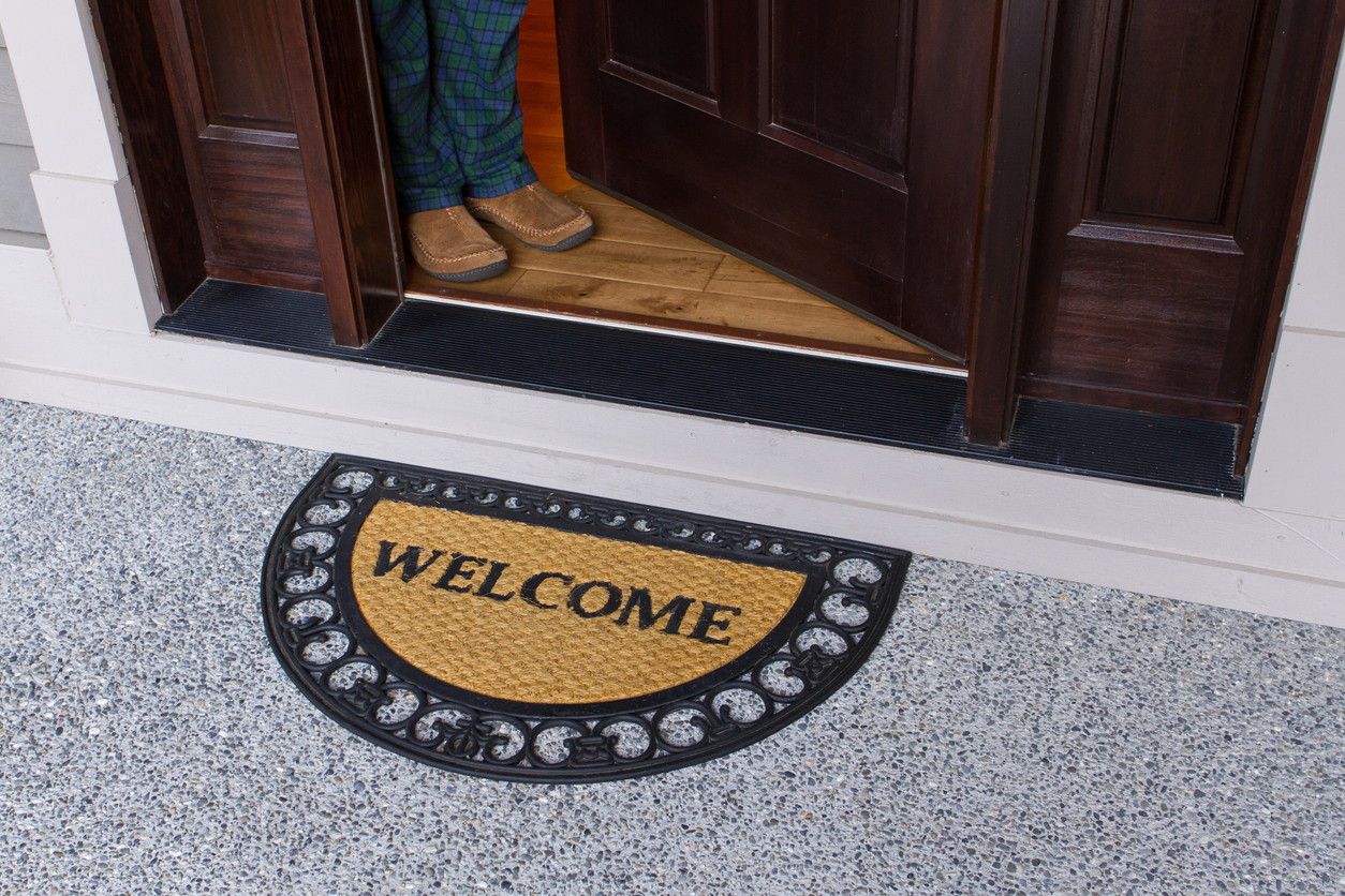 Welcome sign door mat on a porch and open door, person's legs visible on a background.
