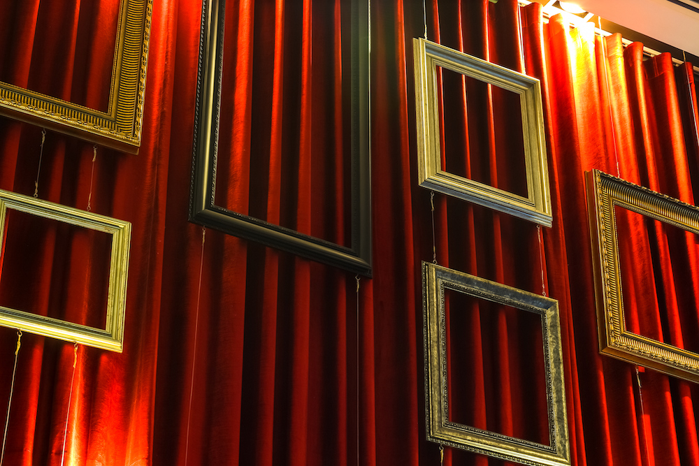 Empty-gold-picture-frames-are-suspended-by-wires-in-front-of-a-red-curtain.