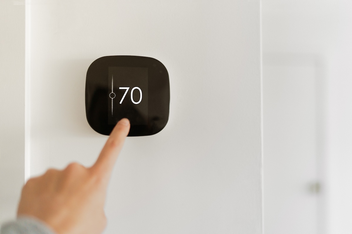 Unseen person uses finger to touch a black smart thermostat set to 70 degrees F on a wall.