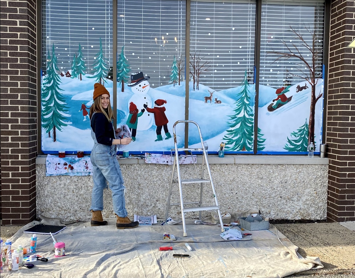 A woman stands next to a wall mural she is painting of a snowy scene outdoors on a window.