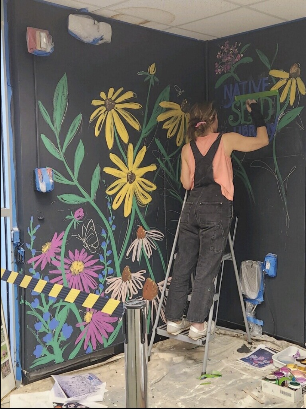 A person stands on a step ladder and paints a colorful floral mural on a black wall.