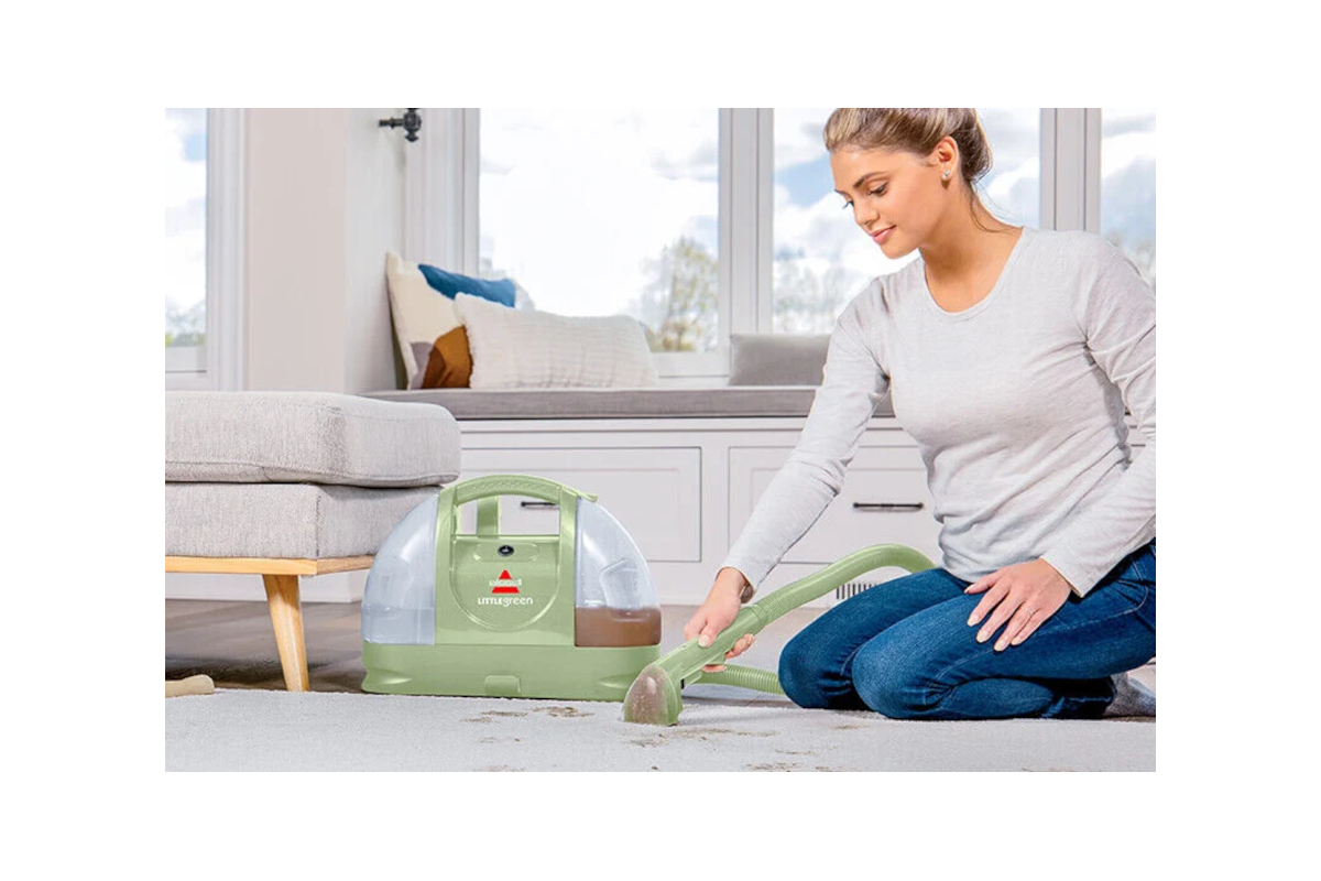 A woman kneeling to clean a heavily stained area rug with a Bissell Little Green portable carpet cleaner.