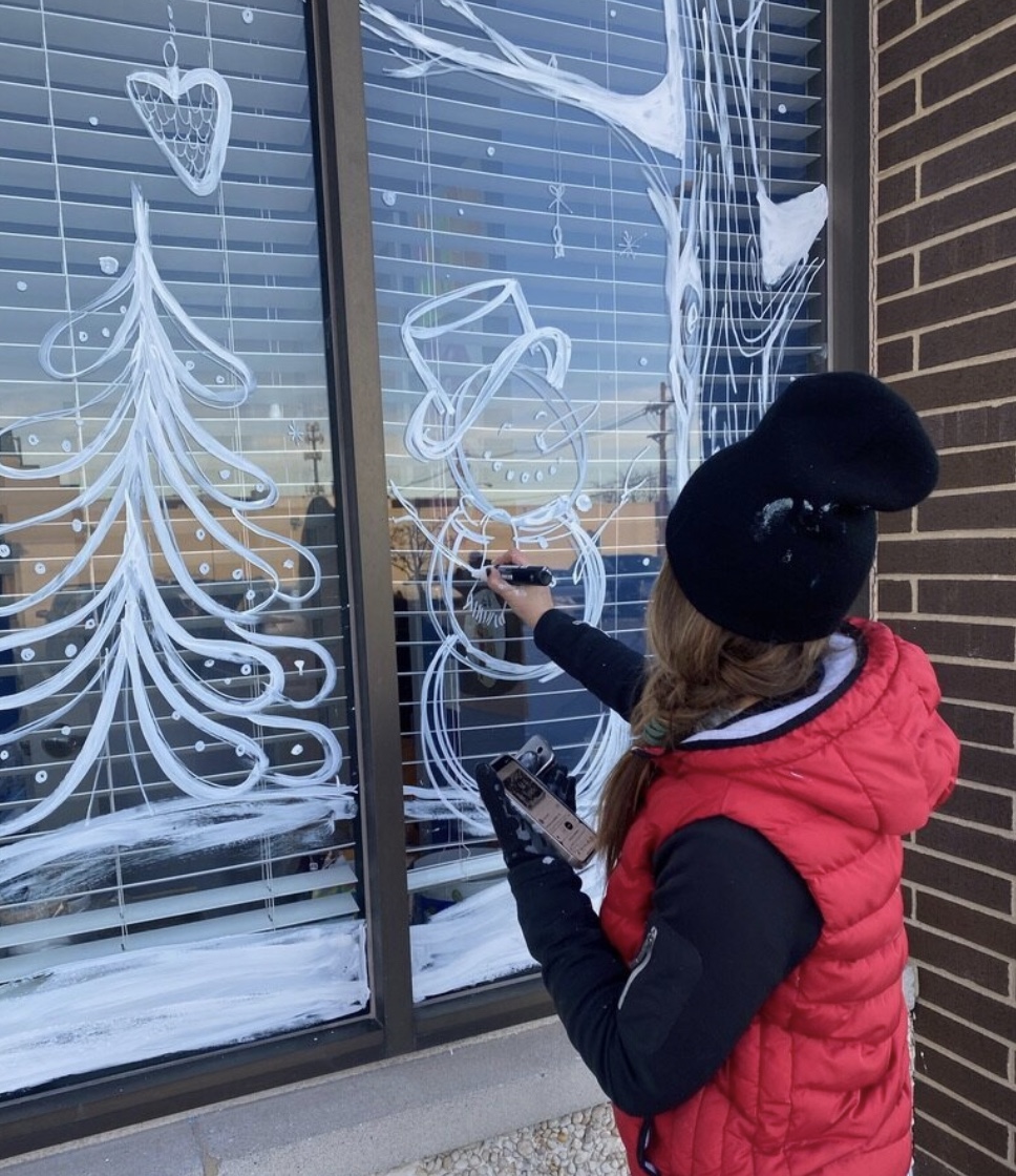 A person uses white paint to sketch out a mural design on a window.