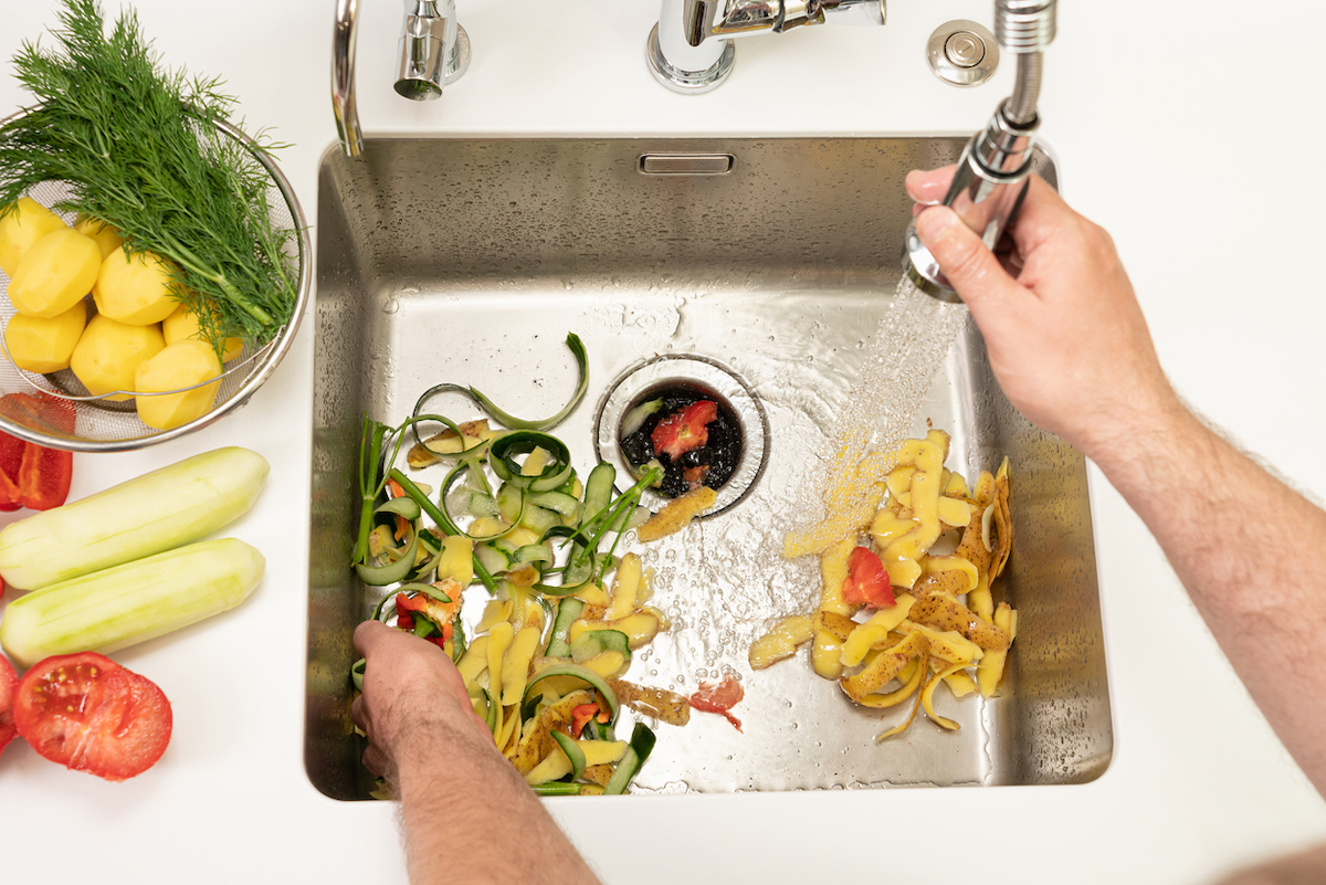 Man uses kitchen sink faucet spray to wash vegetable peels toward kitchen drain.