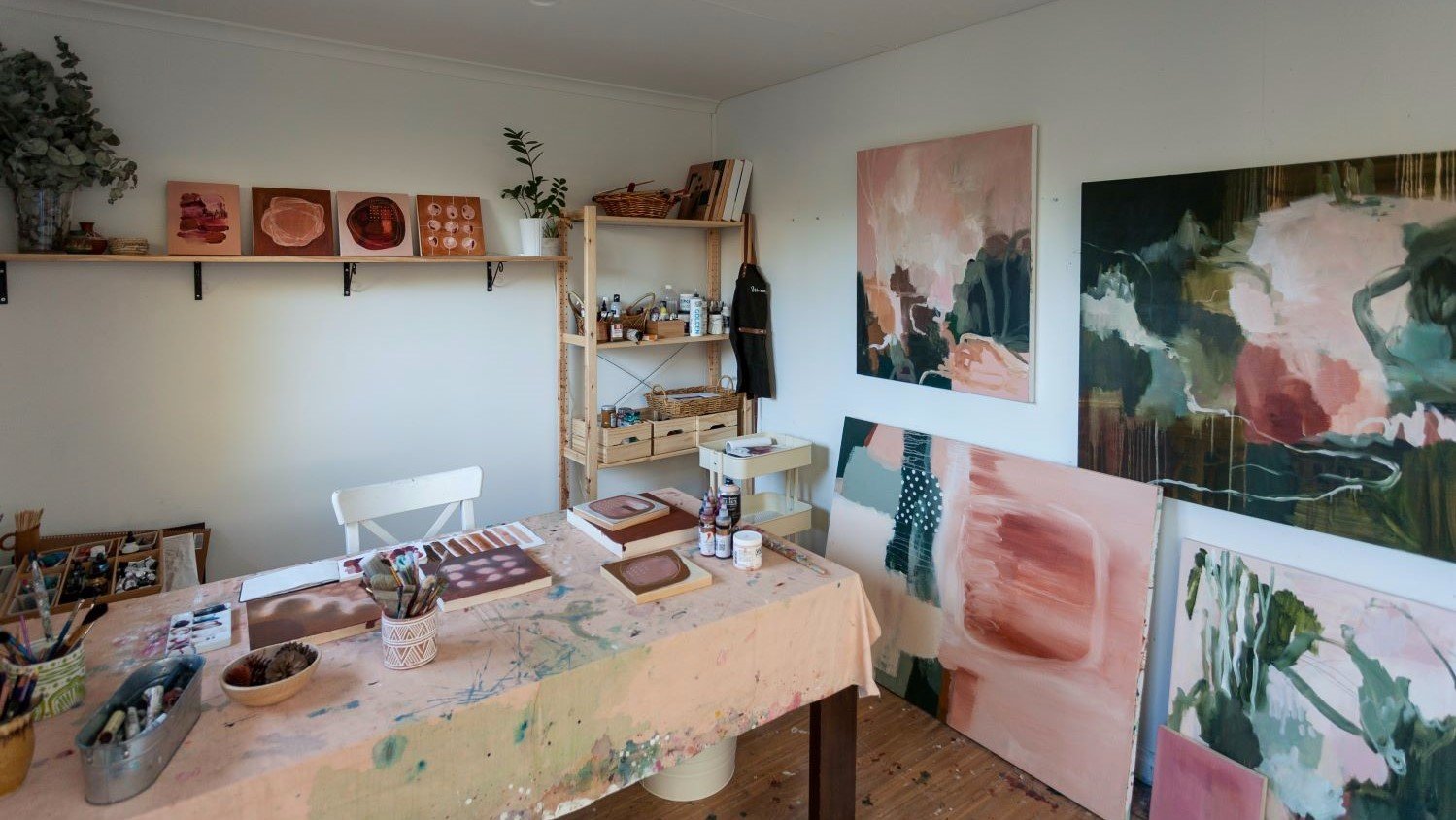 Art studio with green, pink, and beige abstract art on walls above a messy work desk.