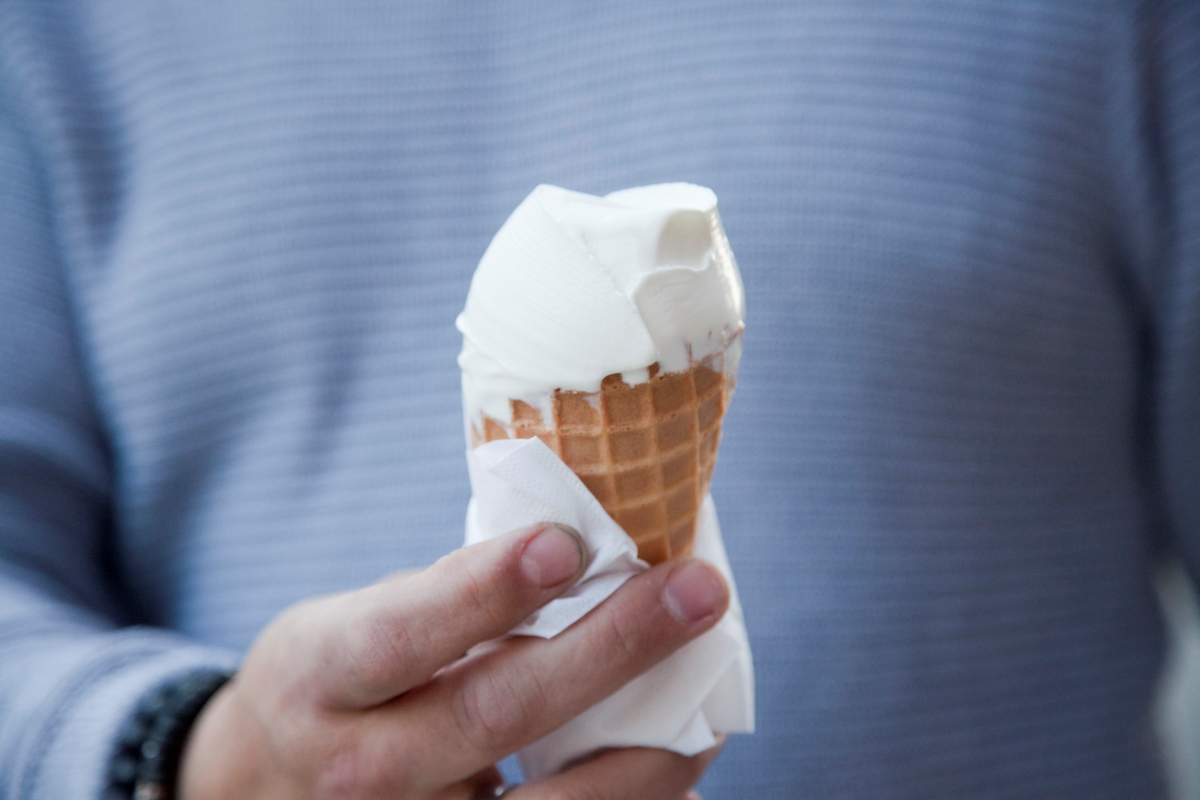 Person in blue shirt holding vanilla ice cream cone wrapped in paper.