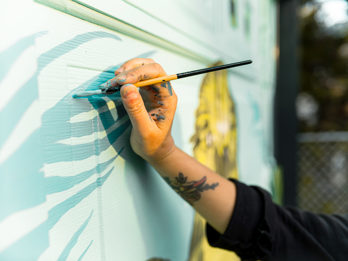 A-hand-with-paint-marks-on-it-holds-a-paint-brush-and-adds-teal-lines-to-a-mural.