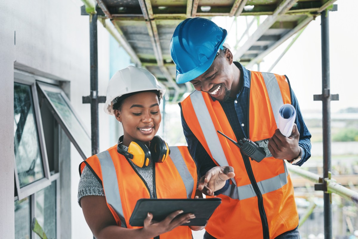 A Black man and woman in construction clothing and hardhats smiling as they overview a project.
