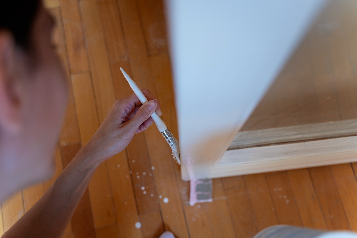 A person painting a wall with paint drips on the hardwood floors underneath.