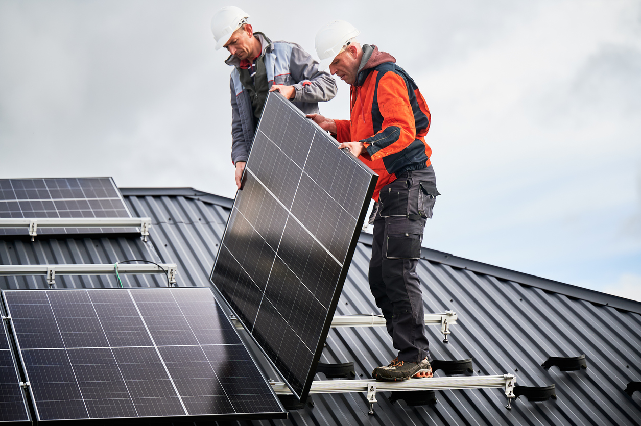 Two men in hardhats install a solar panel on house roof.