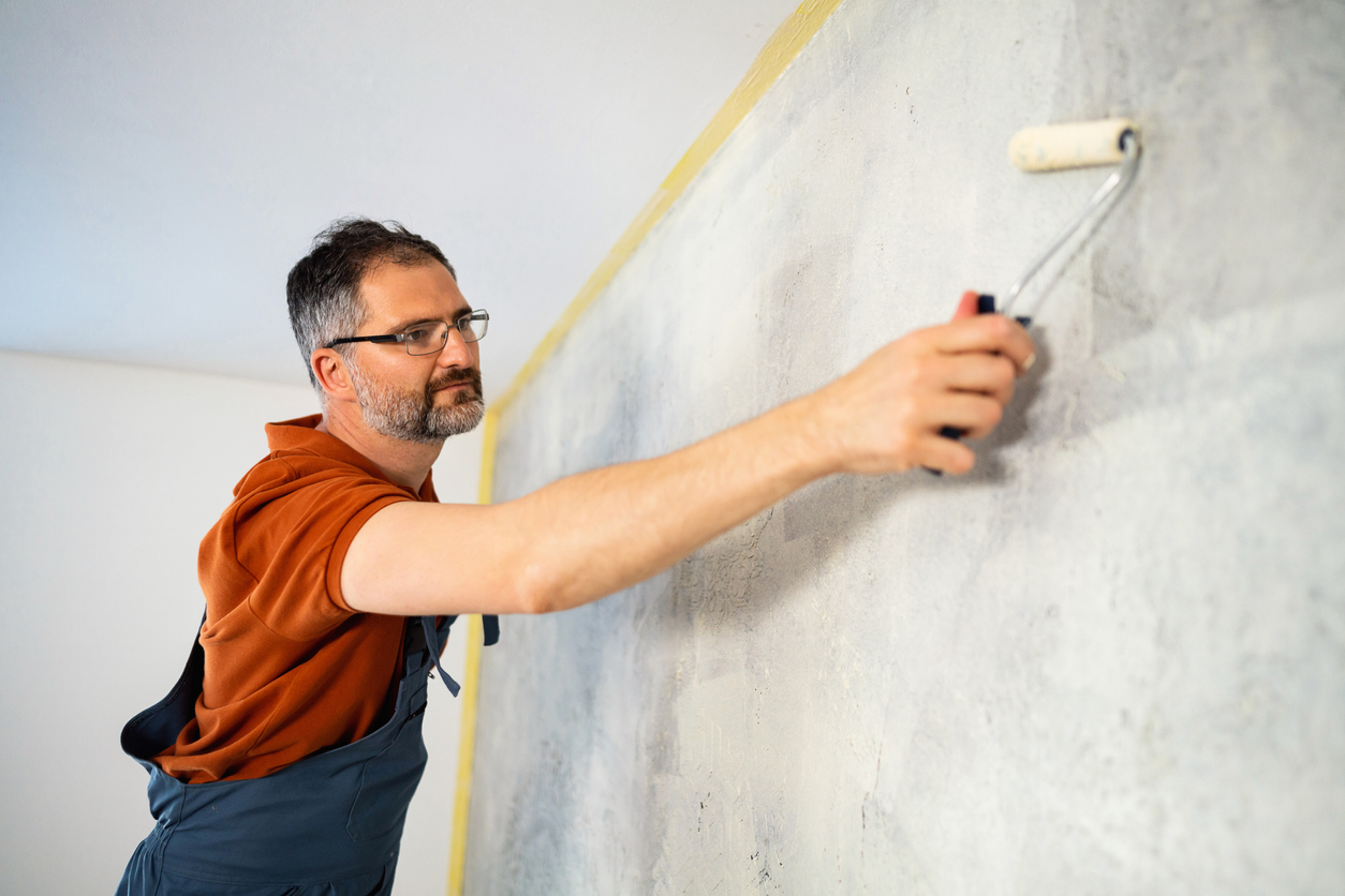 A-man-leans-over-to-reach-part-a-wall-that-he-is-painted-with-a-roller-brush-and-primer.