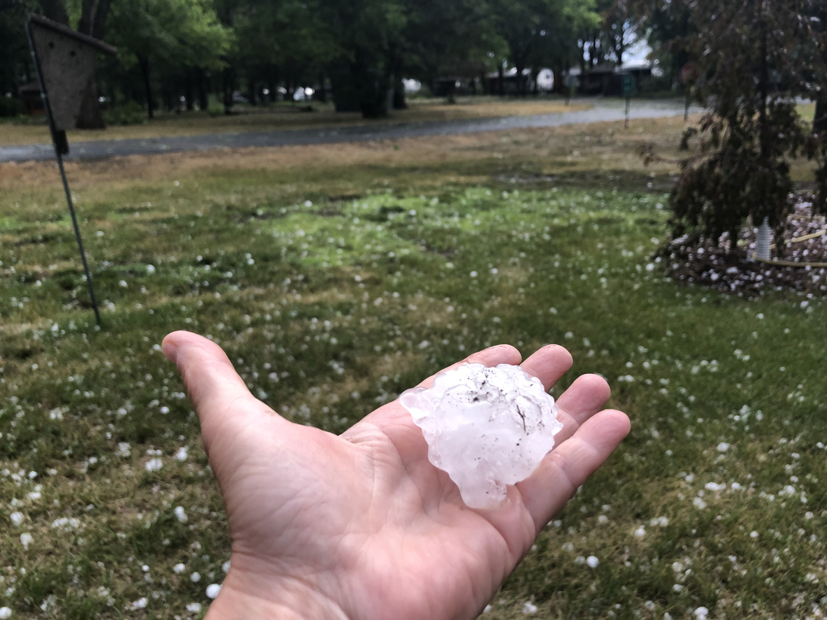A-hand-holds-a-golf-ball-sized-hail-stone-in-front-of-a-grassy-lawn-with-scattered-hail-stones.