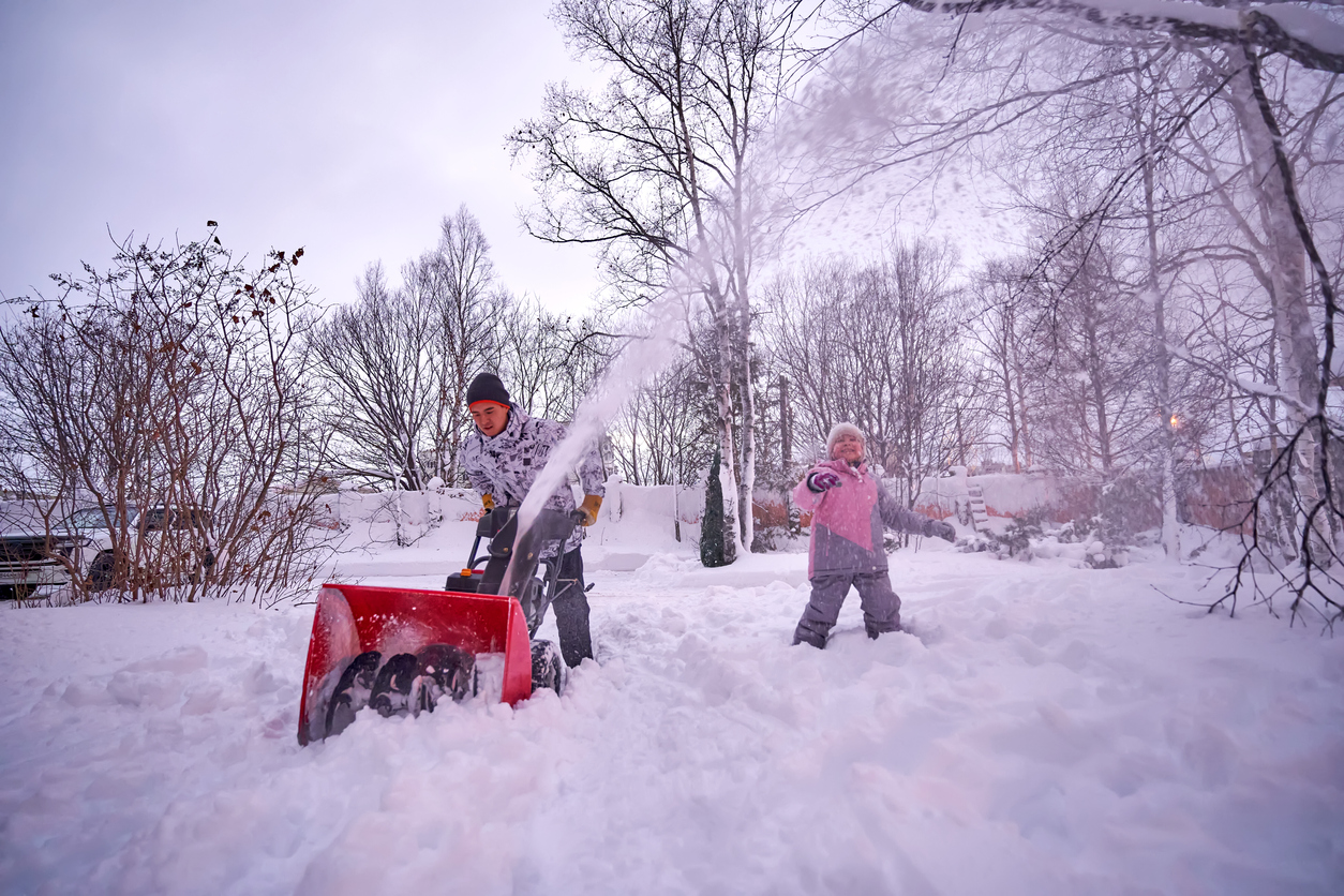 Man in white jacket and hot uses red snowblower while child in pink jacket plays nearby.