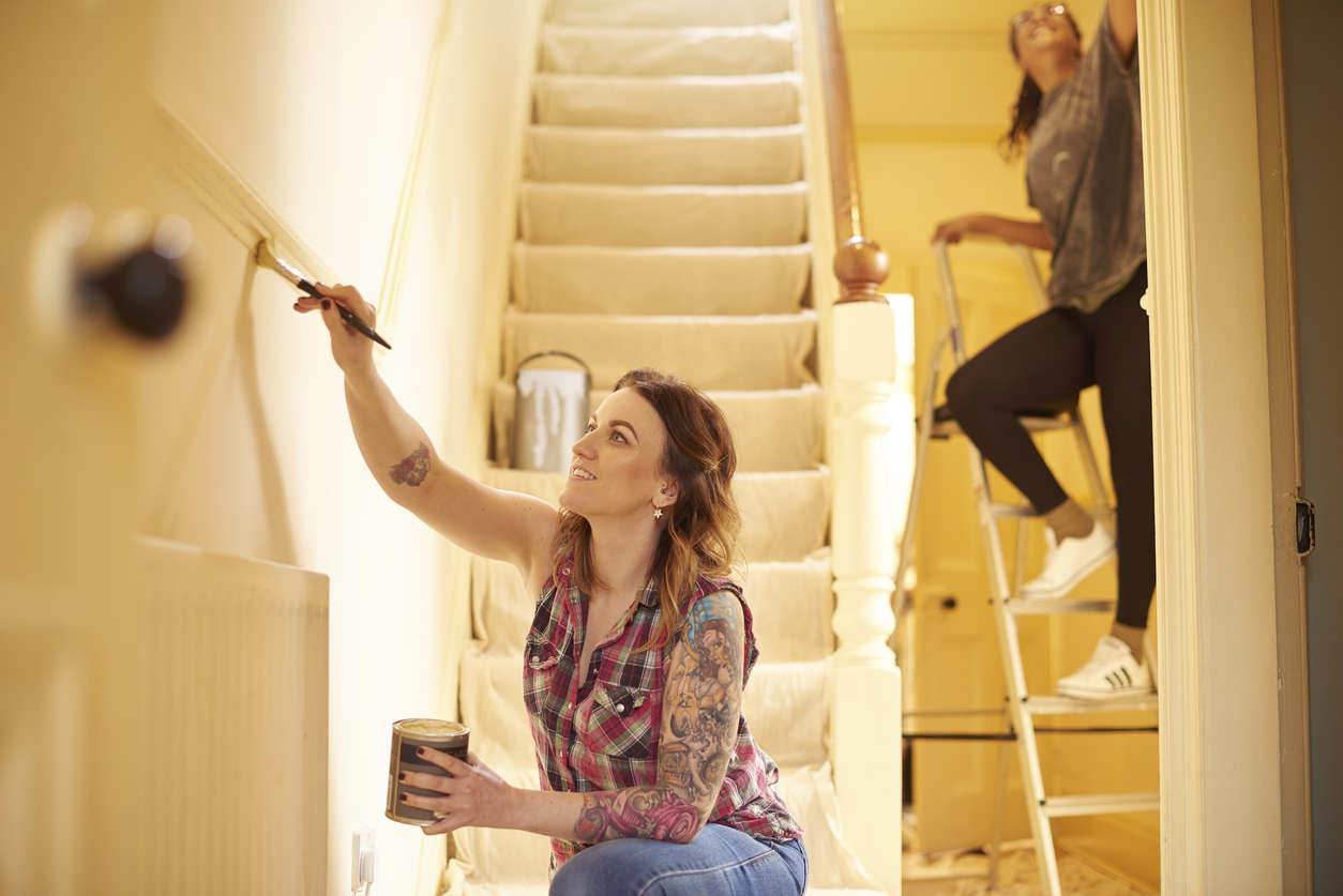 Young woman with tattoos paints staircase with the help up another woman on a ladder.