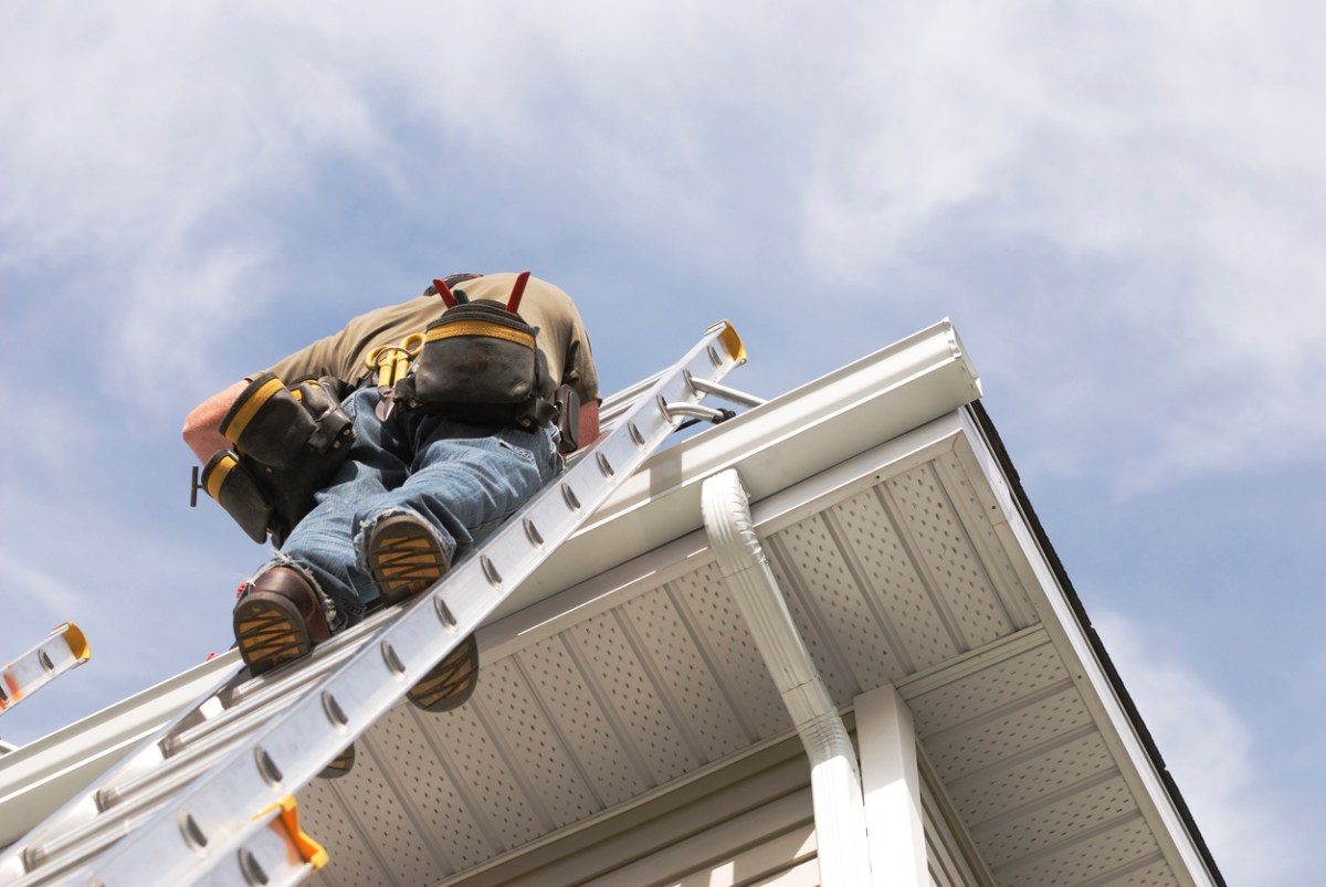 Low angle of a man with tools climbing a ladder onto roof of a house.