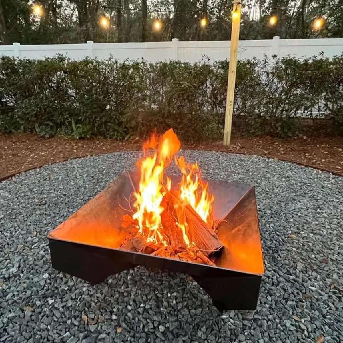 A square metal fire pit with a roaring fire inside, above a gravel base.