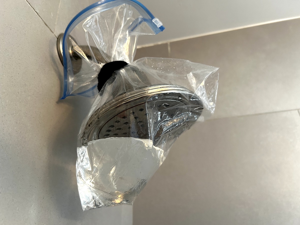 A ziploc bag filled with vinegar tied around a shower head in a grey tiled shower.