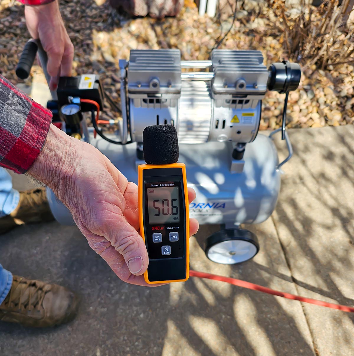 A person holding a decibel meter reading 50.6 next to the California Air Tools 8010 air compressor during testing.