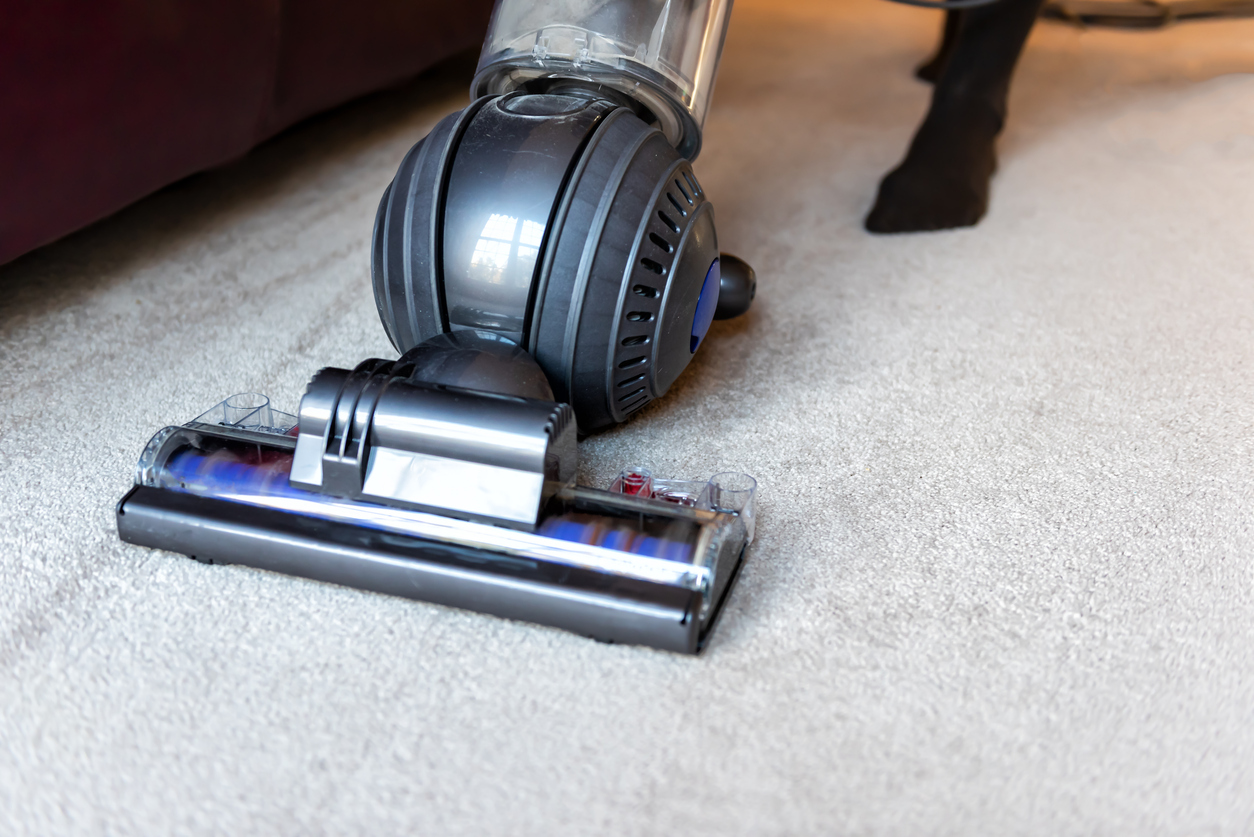 A Dyson Ball standing vacuum cleaner being used on white carpet by a person in black socks.