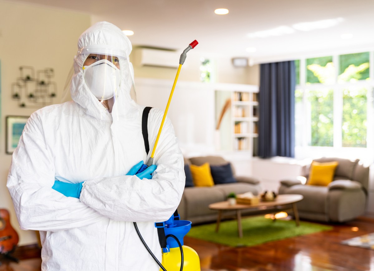 A worker in a full white suit poses in a living room with a pest control tool.