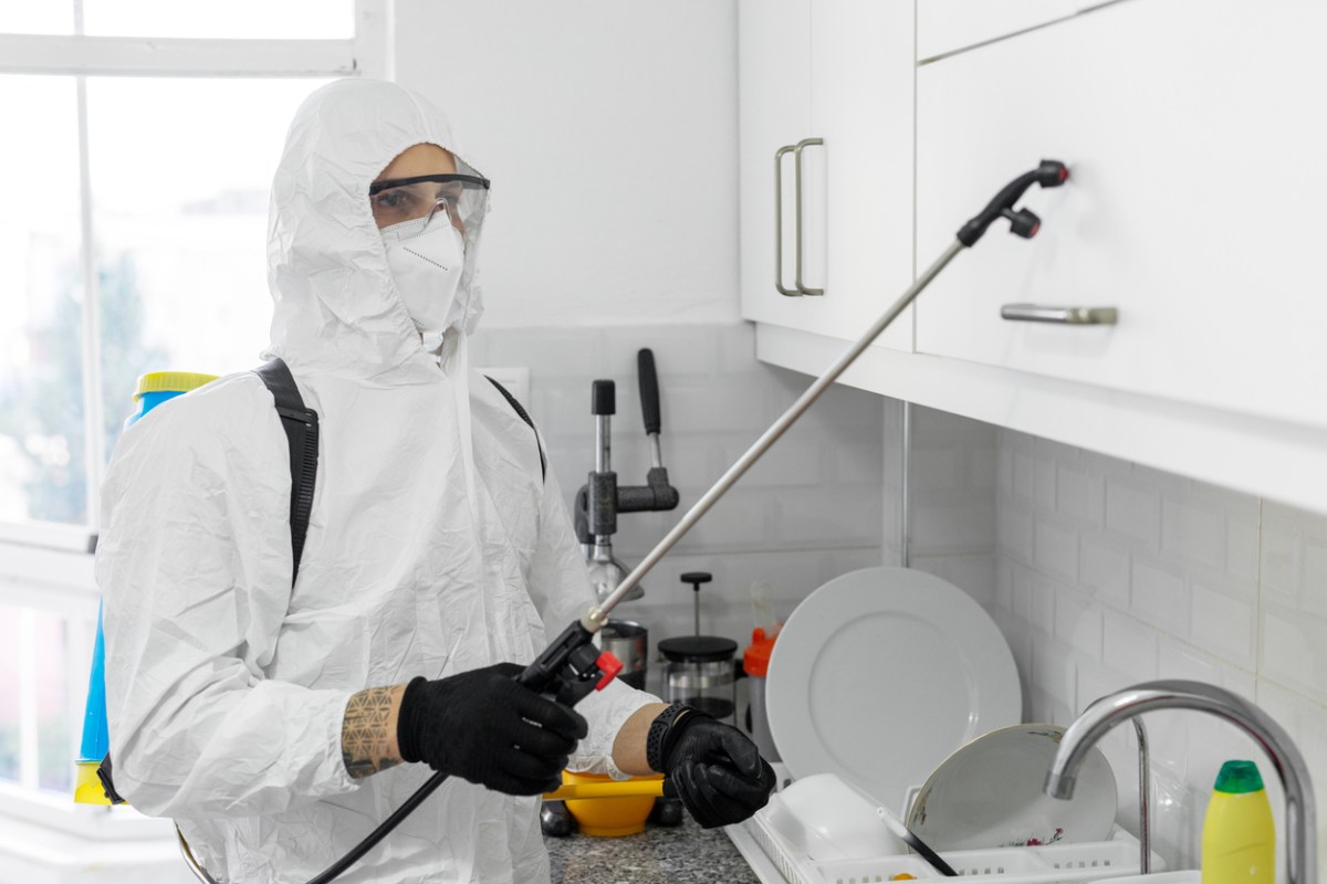 A person in a hazmat suit uses a tool to spray a pest control solution in a kitchen.