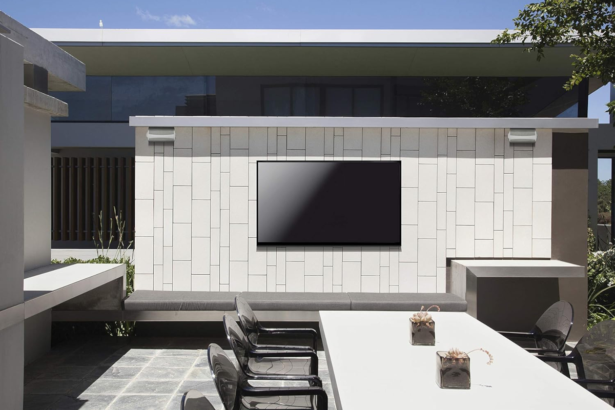 A modern backyard patio area with a large TV and two speakers mounted on an exterior home wall.