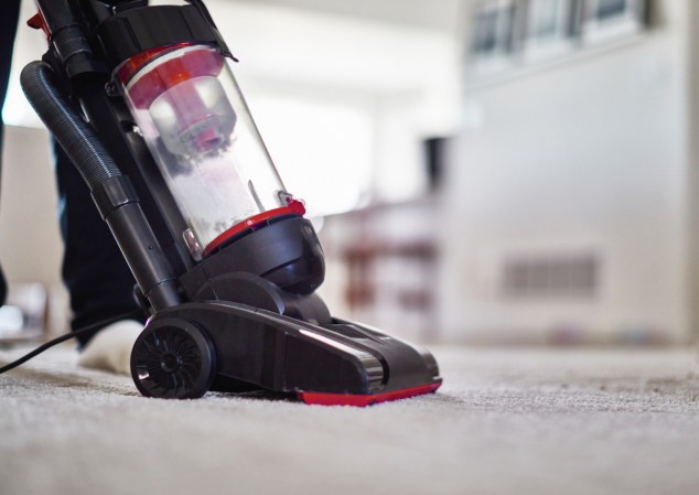 Light carpet being cleaned with a black-and-red standing vacuum cleaner with a bagless dust compartment.