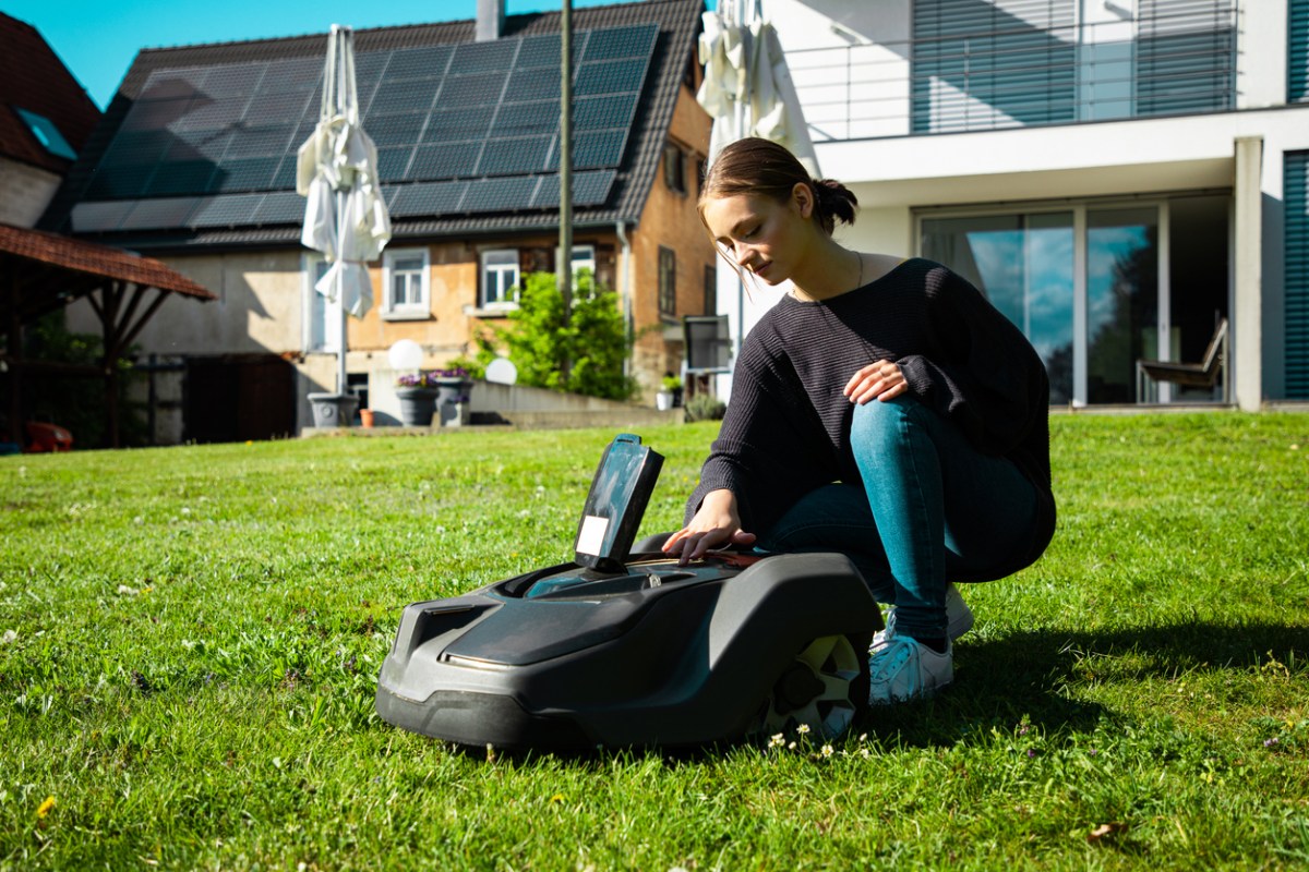 A young woman accessing a control panel on the top of a robot lawn mower in the yard of her upscale smart home.