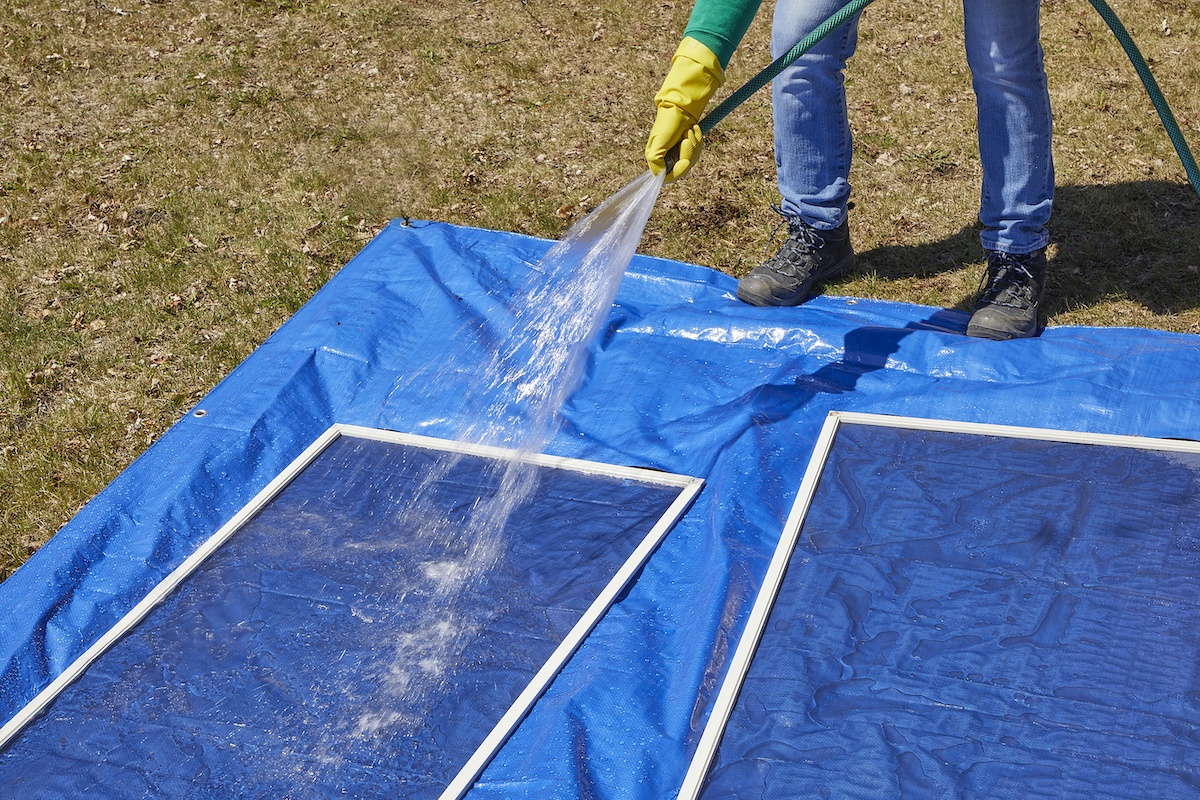 Woman rinsing two window screens on top of a blue tarp outside.