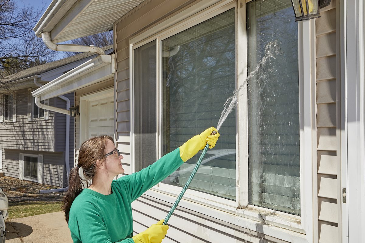 Woman uses a garden hose to rinse exterior windows.