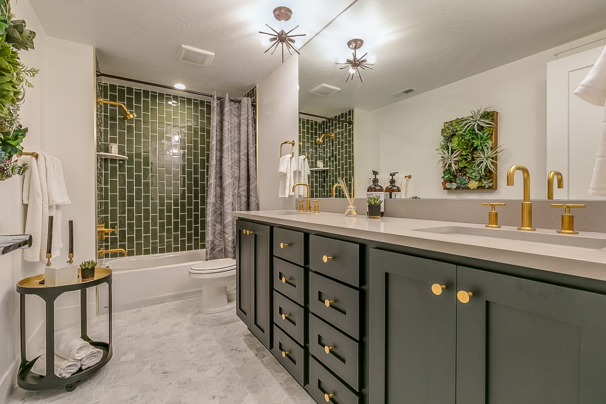 Home bathroom with green tile shower, gold fixtures, a dark green double vanity, and house plants.