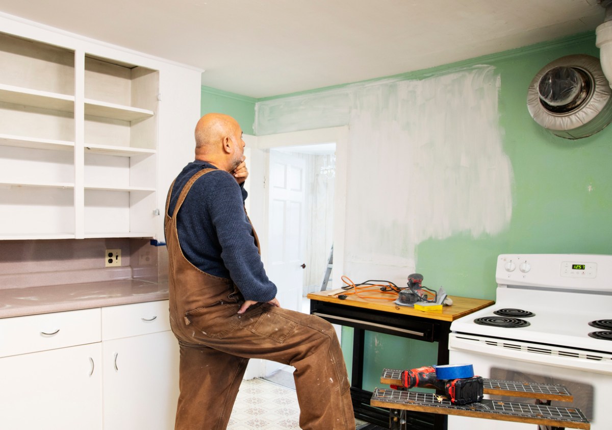 A-man-in-overalls-considers-a-partially-painted-wall-in-a-small-mismatched-kitchen.