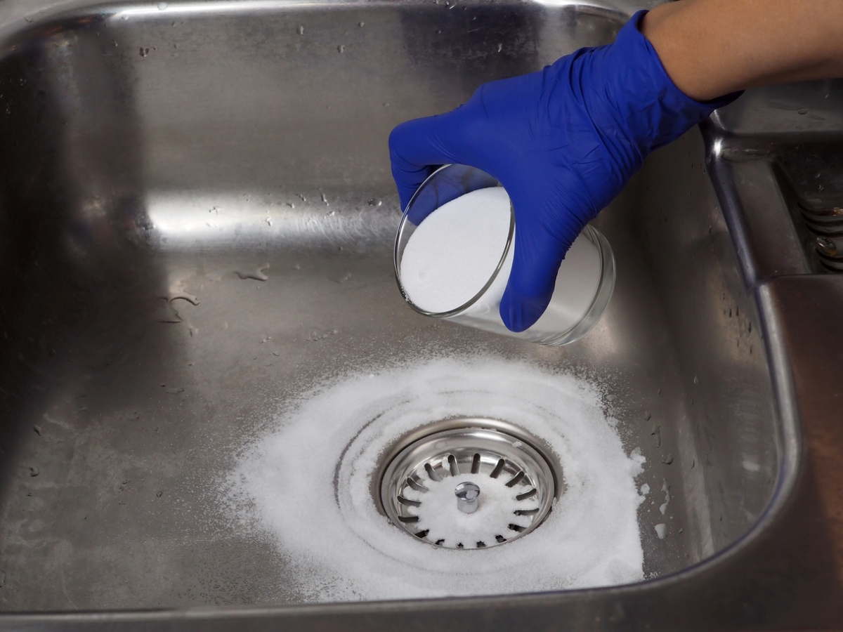 Gloved hand pouring baking soda from glass in ktichen drain.