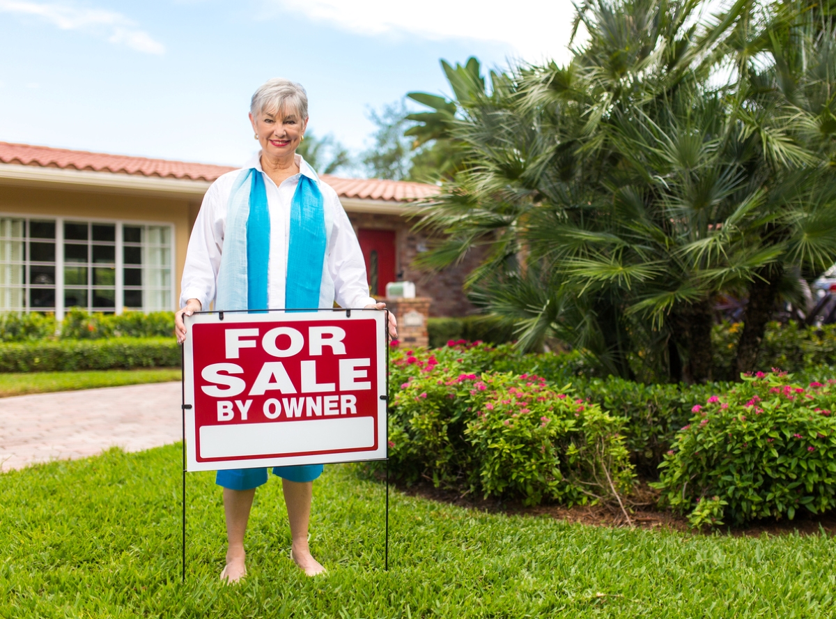 Older woman with for sale by owner sign in yard.