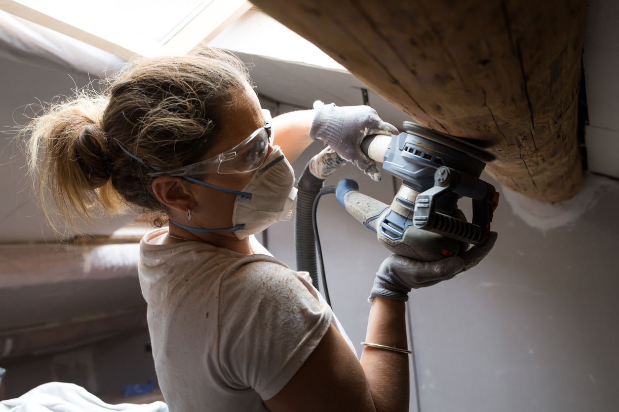 A-woman-uses-a-sander-on-a-wooden-beam-while-wearing-a-mask.
