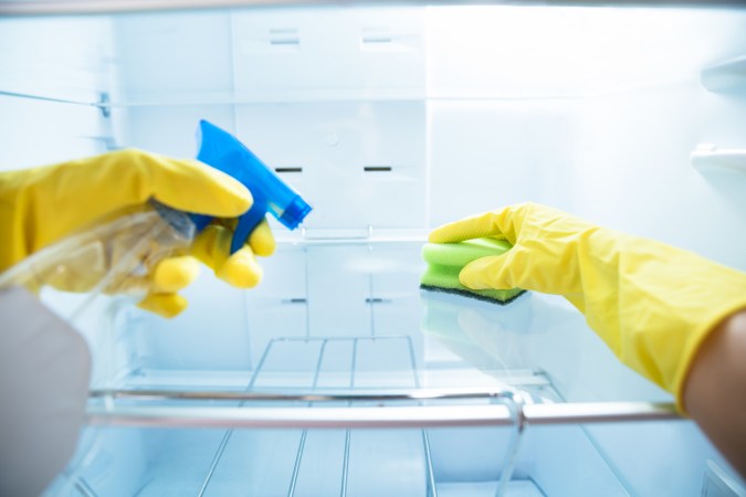 A-persons-hands-in-yellow-gloves-use-spray-cleaner-and-a-magnetic-sponge-to-clean-refrigerator-shelves.