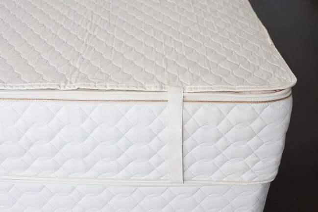 A white cotton quilted mattress pad is secured to a white mattress with a strap on the corner.
