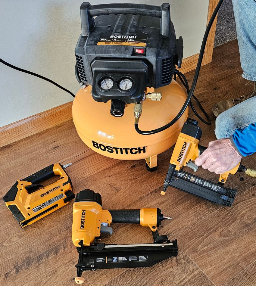 A person holding a Bostitch pneumatic tool that's connected to the Bostitch pancake air compressor with two other Bostitch tools next to it.