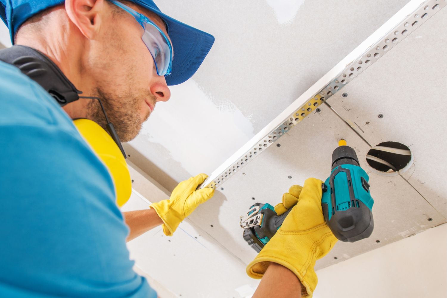 A worker in yellow gloves uses a drill to install drywall.